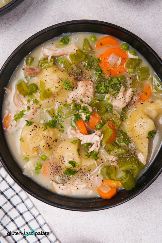 A black bowl filled with gluten-free dumplings, shredded chicken, carrots, celery, peas, and broth, topped with chopped parsley and black pepper.