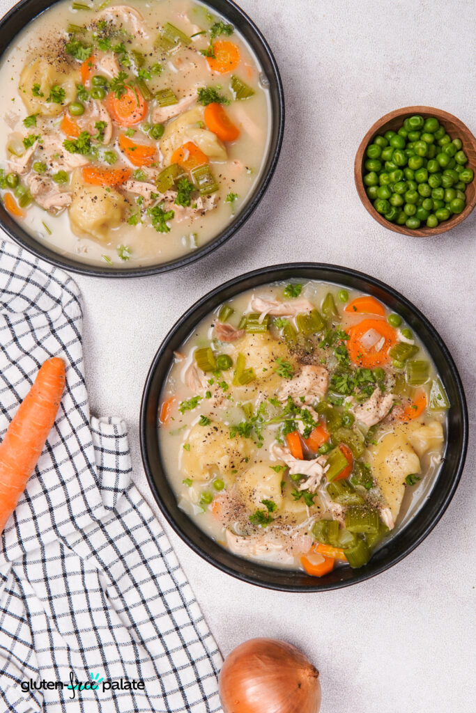 Top-down view of bowls containing gluten-free dumplings, chicken, celery, carrots, peas, and broth, with parsley sprinkled on top and a bowl of peas on the side.