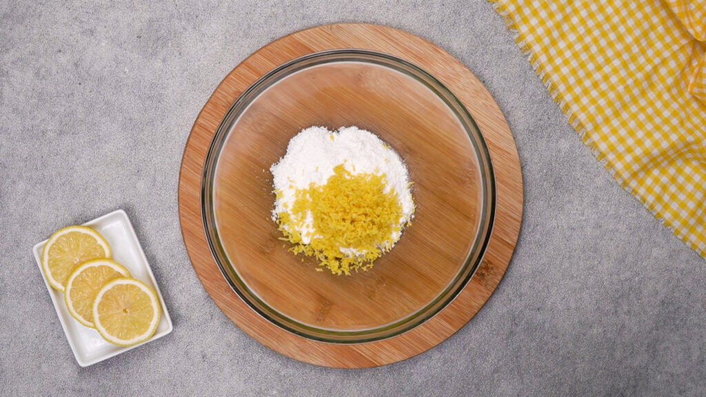 A glass mixing bowl on a wooden board filled with powdered sugar and fresh lemon zest, ready to be rubbed together for the lemon crinkle cookie dough.