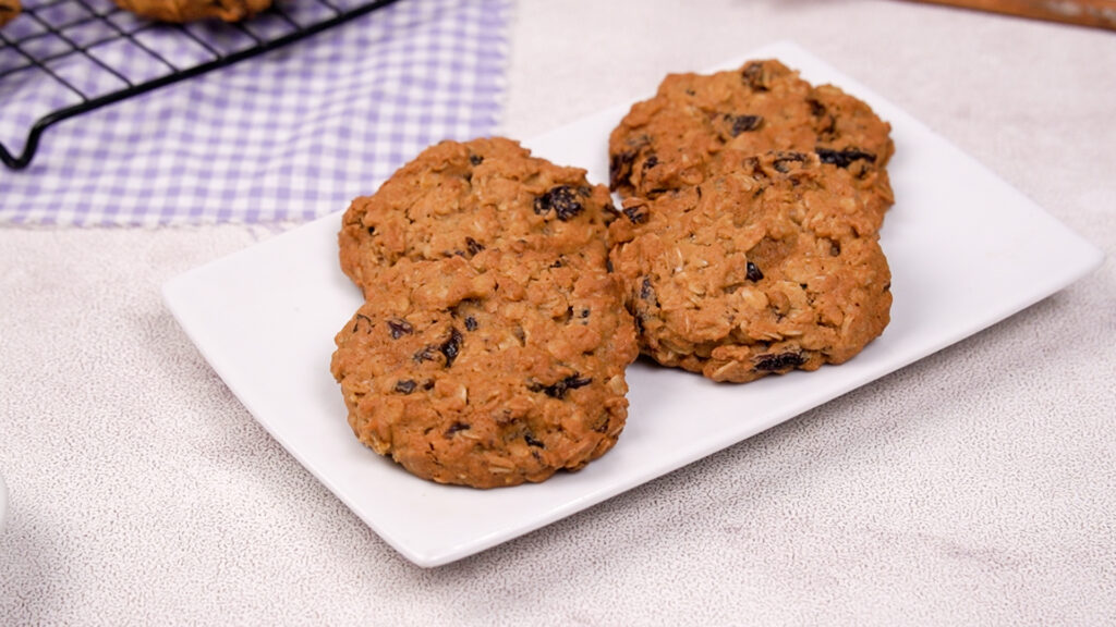 A close-up of four finished cookies on a white plate, showcasing their texture and raisins.