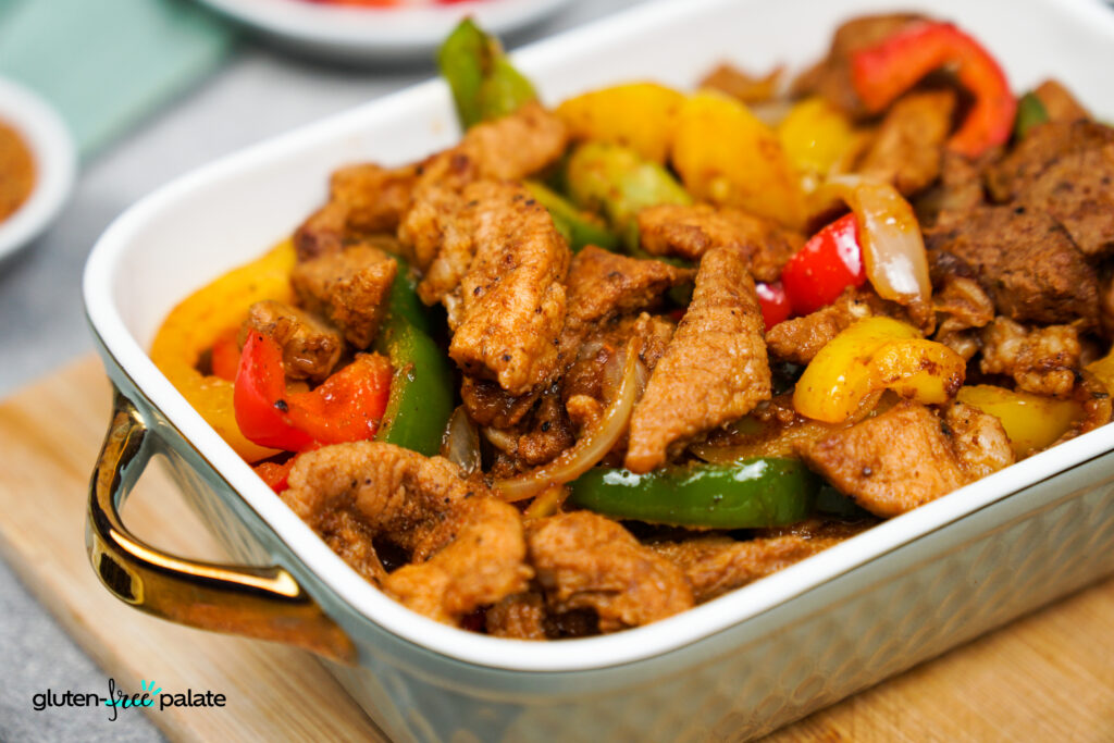 Rectangular white ceramic baking dish overflowing with cooked strips of seasoned pork, sliced red, yellow, and green bell peppers, and onions.