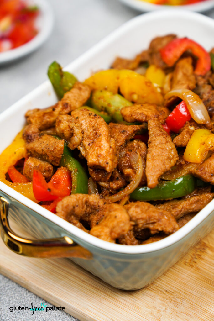 Tight shot of juicy pork fajita strips mixed with bright red, yellow, and green pepper slices and translucent onion in a white serving dish.