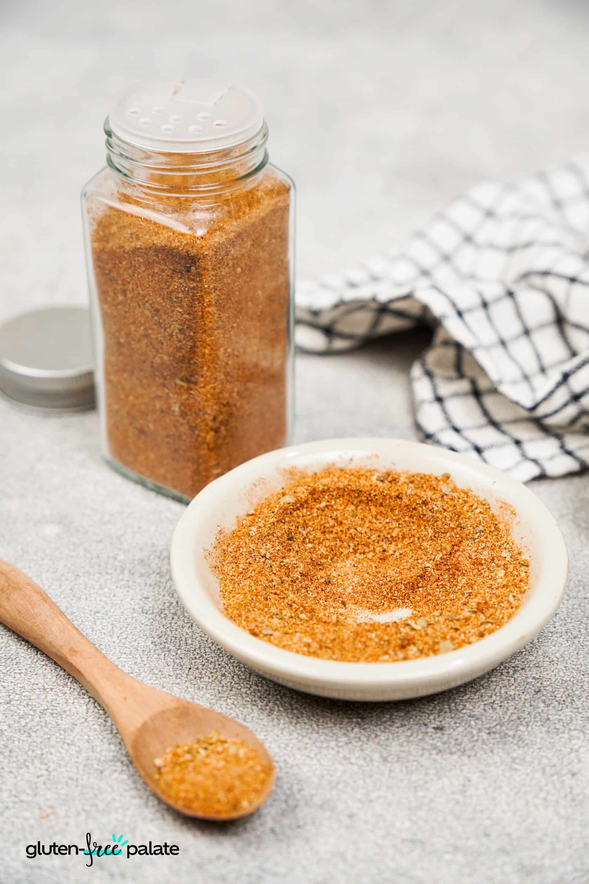 Glass spice jar and a small white bowl filled with the same orange-brown fajita seasoning blend, with a black-and-white checkered cloth in the background.