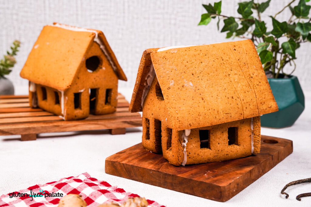 Two gluten-free gingerbread houses displayed on wooden boards, decorated with royal icing and surrounded by plants and holiday decor.