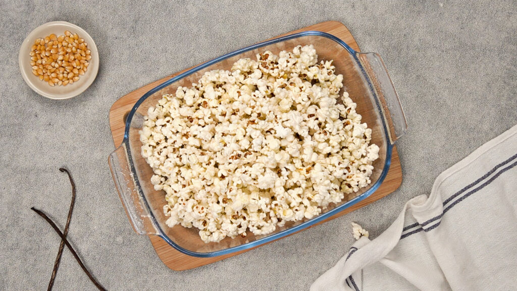 A clear rectangular baking dish filled with freshly popped popcorn sits on a wooden board.