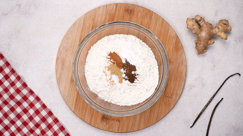 Bowl with gluten-free flour, ginger, cinnamon, nutmeg, cloves, baking soda, and salt mixture on a wooden board.