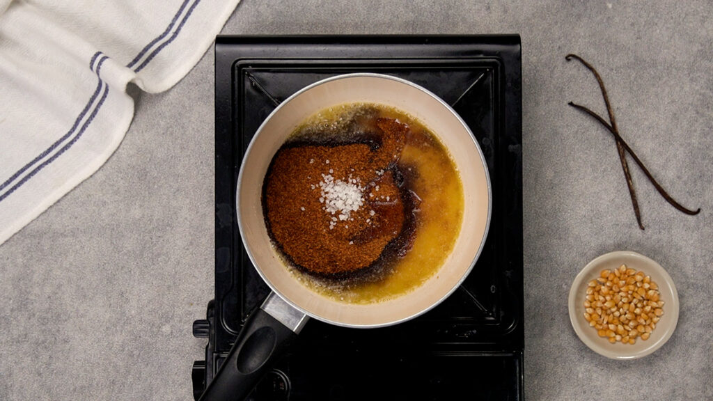 Top-down view of a saucepan on a portable stovetop burner. Melted butter, brown sugar, corn syrup, and a sprinkle of coarse sea salt sit unmixed in the pan.