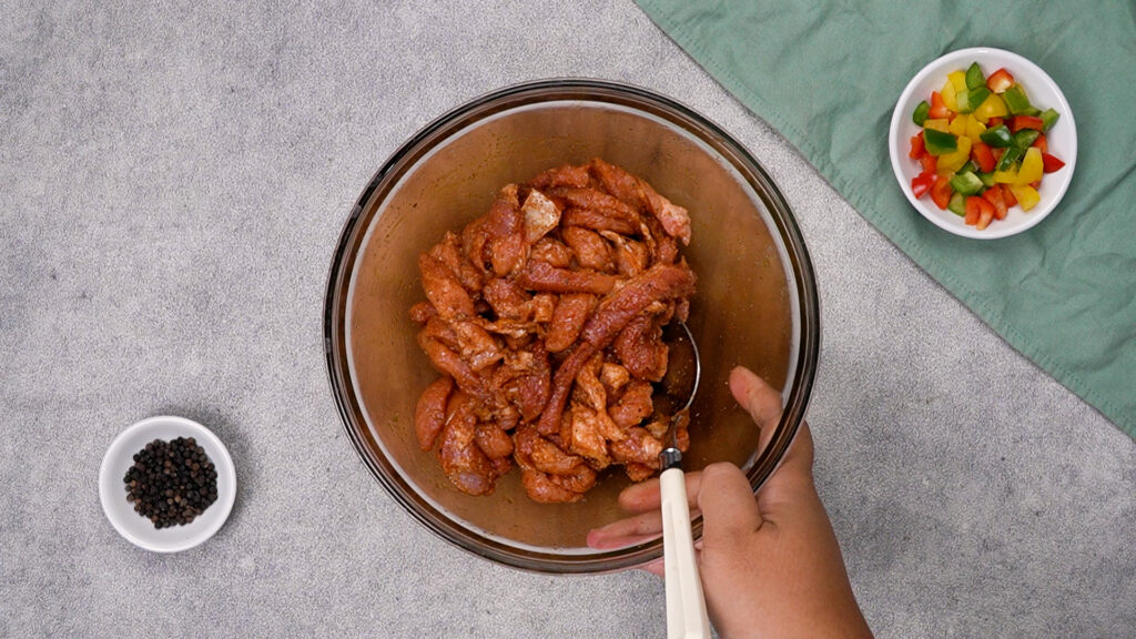 A glass mixing bowl filled with raw pork bites coated in fajita spice blend, being stirred with a spoon.