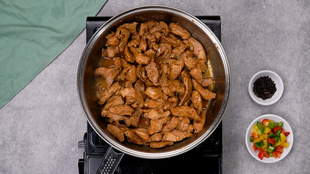 A stainless-steel skillet on a portable burner holding browned, cooked pork strips.