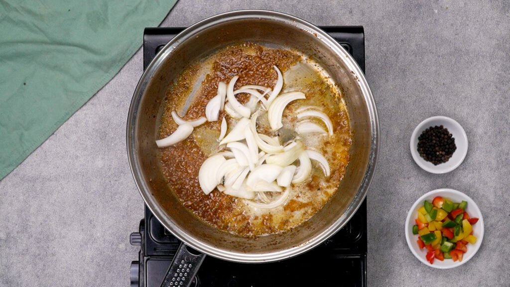 Overhead shot of sliced white onions being added to a skillet with residual cooking oil and spice bits.