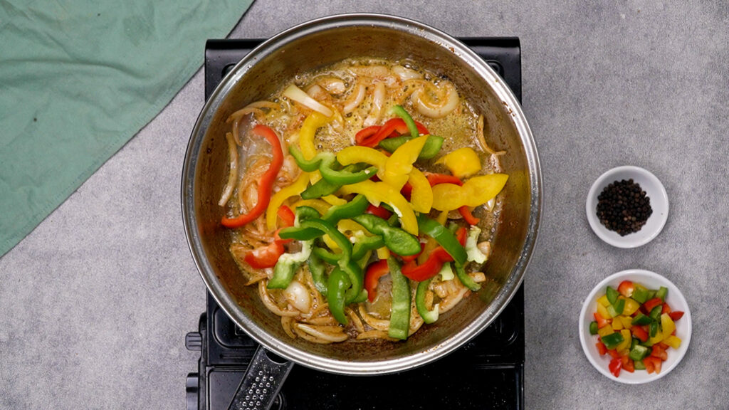 Overhead view into a hot skillet where sliced onions and red, yellow, and green bell pepper strips are sautéing in oil.
