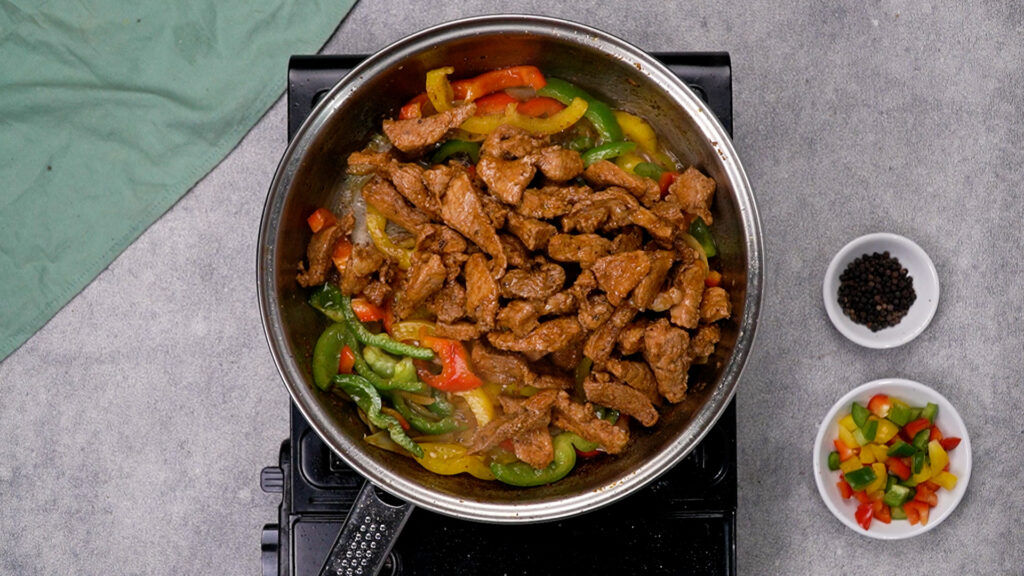 Overhead shot of the skillet with pork strips just returned to the pan, nestled into the sizzling peppers and onions.