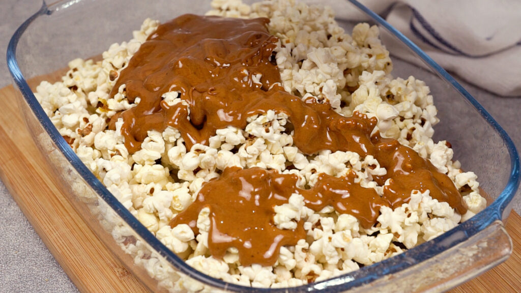 Thick caramel is being poured from the saucepan onto the popcorn in the baking dish, coating the top layer in a rich amber stream.