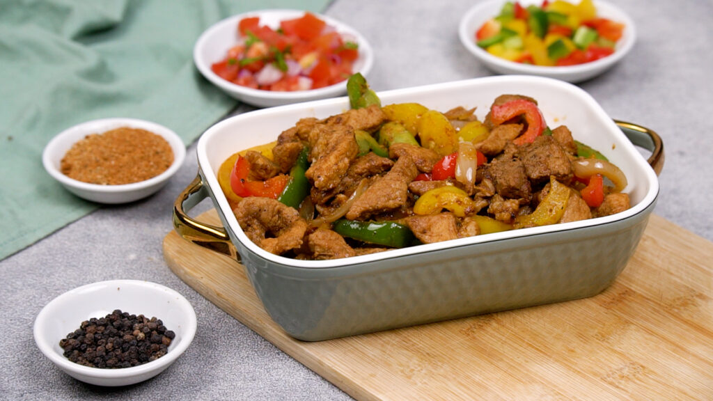 Overhead view of a white rectangular baking dish filled with cooked, seasoned pork fajita strips and sautéed red, yellow, and green peppers and onions, set on a wooden board.
