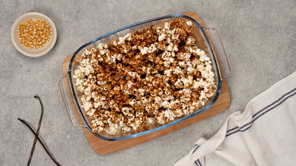 Partially coated popcorn in the dish shows some clusters already smothered in caramel while the rest remains white, awaiting mixing.