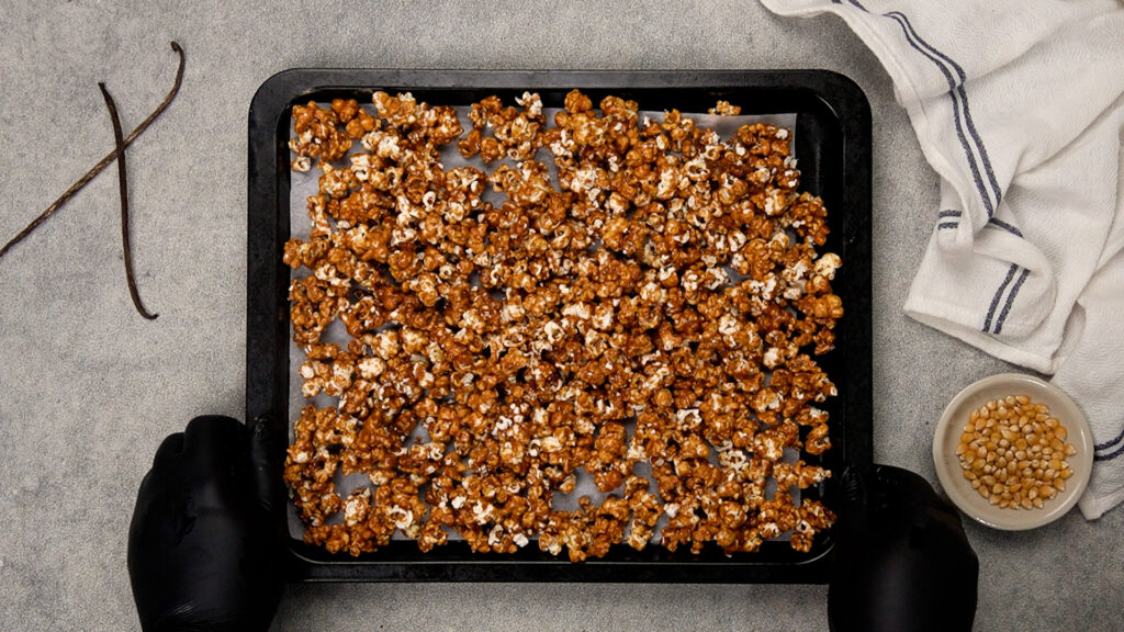 A parchment-lined baking sheet holds caramel-coated popcorn clusters spread out evenly; a hand in a black glove grips the pan’s edge.