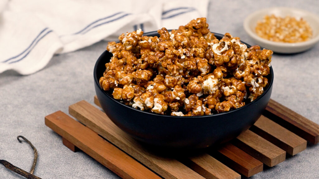 Finished caramel corn fully coated in glossy toffee clusters is heaped in a black serving bowl, with raw kernels and vanilla pods in the background.