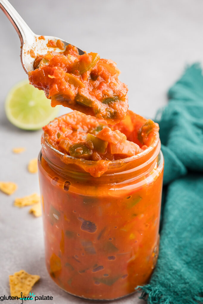 Spoonful of chunky cooked bell pepper salsa being lifted from a glass jar, showing its thick texture and vibrant red-orange color.