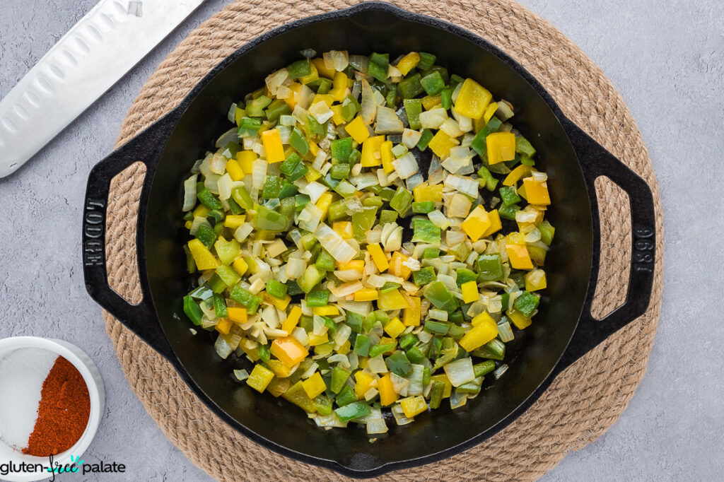 Diced green and yellow bell peppers with onion cooking in a black cast iron pan, next to a knife and seasoning.