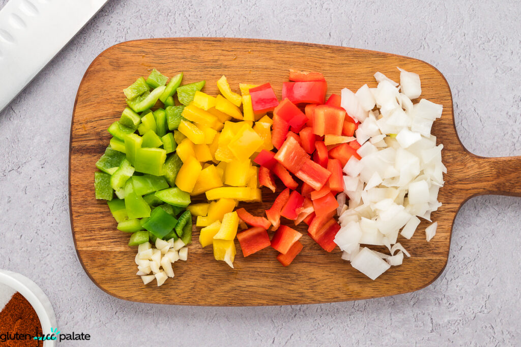 Diced green, yellow, and red bell peppers lined up with chopped white onion and garlic on a wooden cutting board, ready for salsa prep.