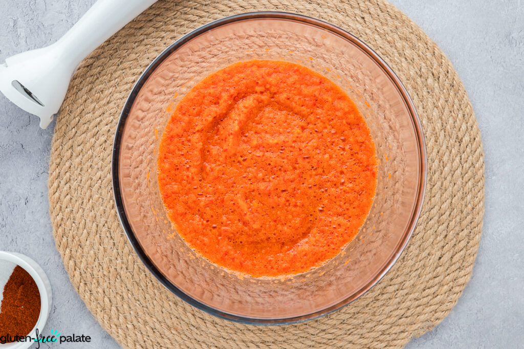 Bright orange-red blended bell pepper mixture in a glass bowl with an immersion blender on the side.