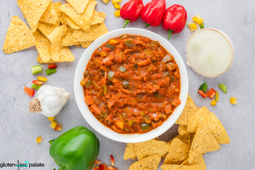 Bowl of gluten-free cooked salsa surrounded by tortilla chips, garlic, bell pepper pieces, and habanero peppers.