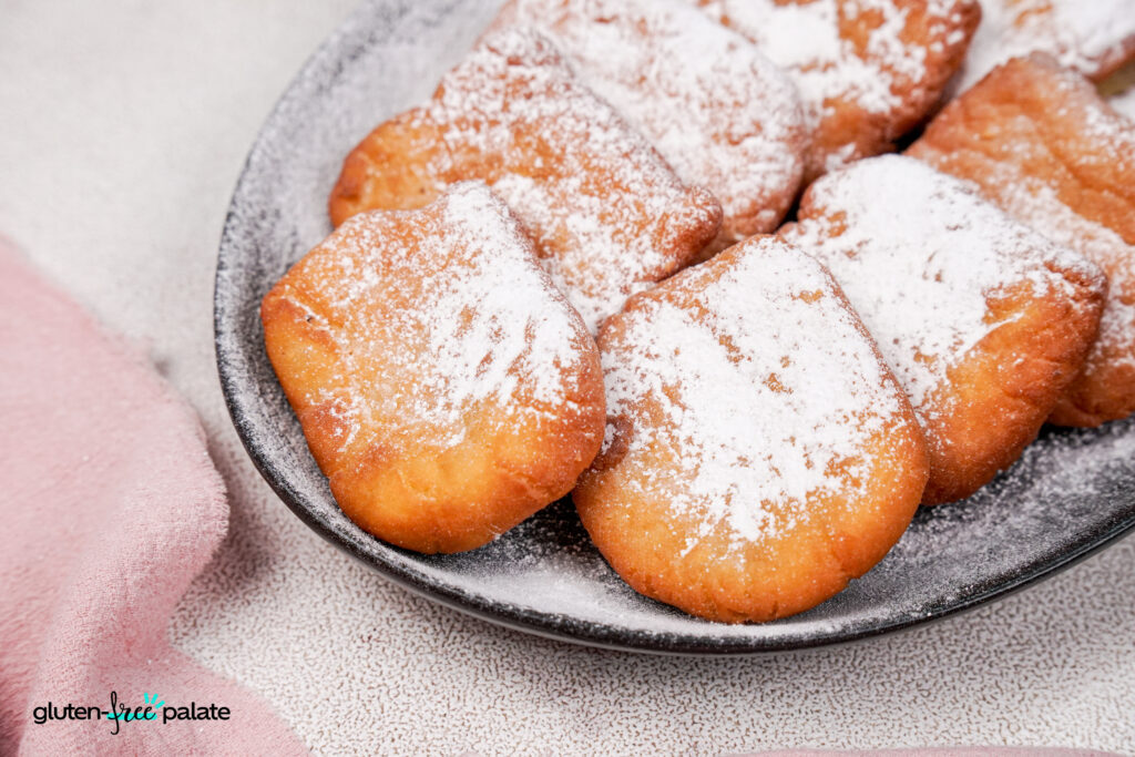 A plate of golden-brown gluten-free beignets generously dusted with powdered sugar, served on a dark plate against a light background.
