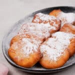 Freshly fried gluten-free beignets on a black plate, topped with powdered sugar, photographed at an angle to show their fluffy texture.