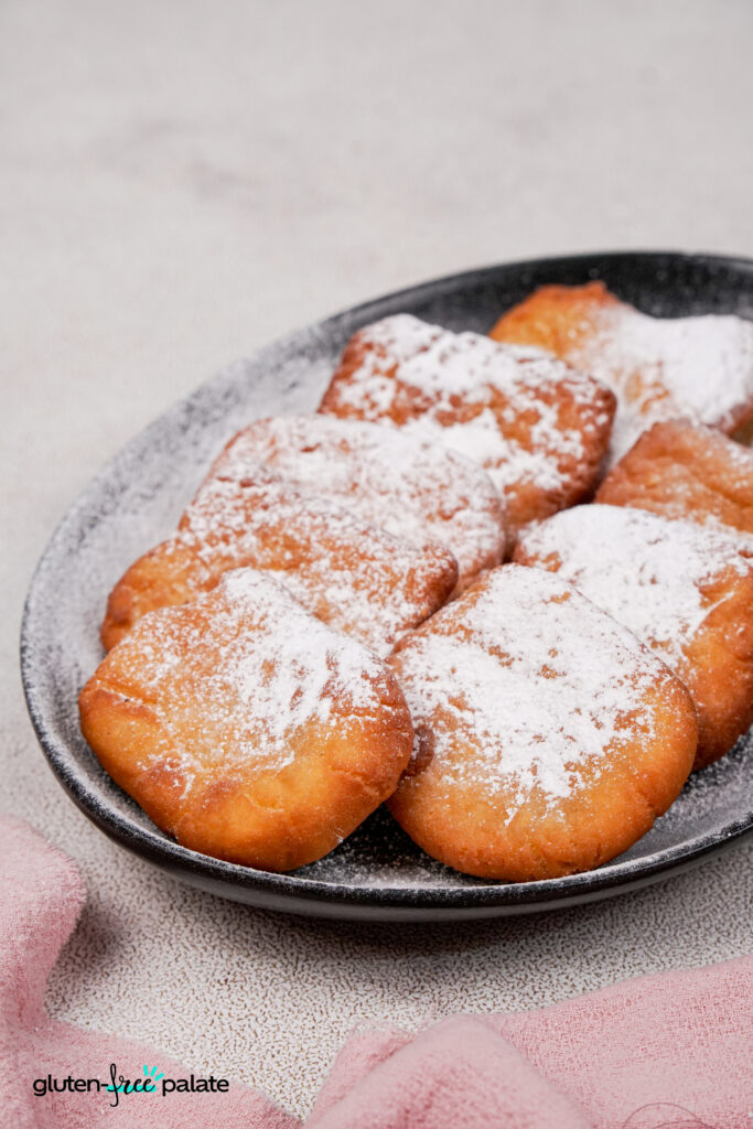 Freshly fried gluten-free beignets on a black plate, topped with powdered sugar, photographed at an angle to show their fluffy texture.