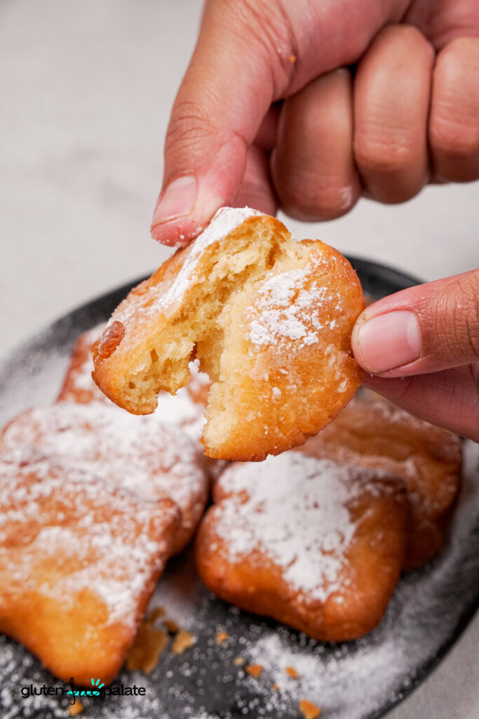 Hands breaking open a warm gluten-free beignet to reveal its soft, fluffy inside, dusted with powdered sugar.