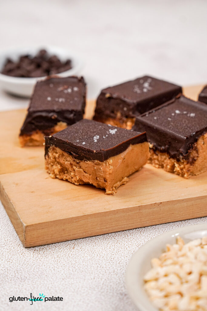 Close-up of a single gluten-free peanut butter bar with a dense peanut butter base and glossy chocolate topping, sprinkled with flaky sea salt, sitting on a wooden board.