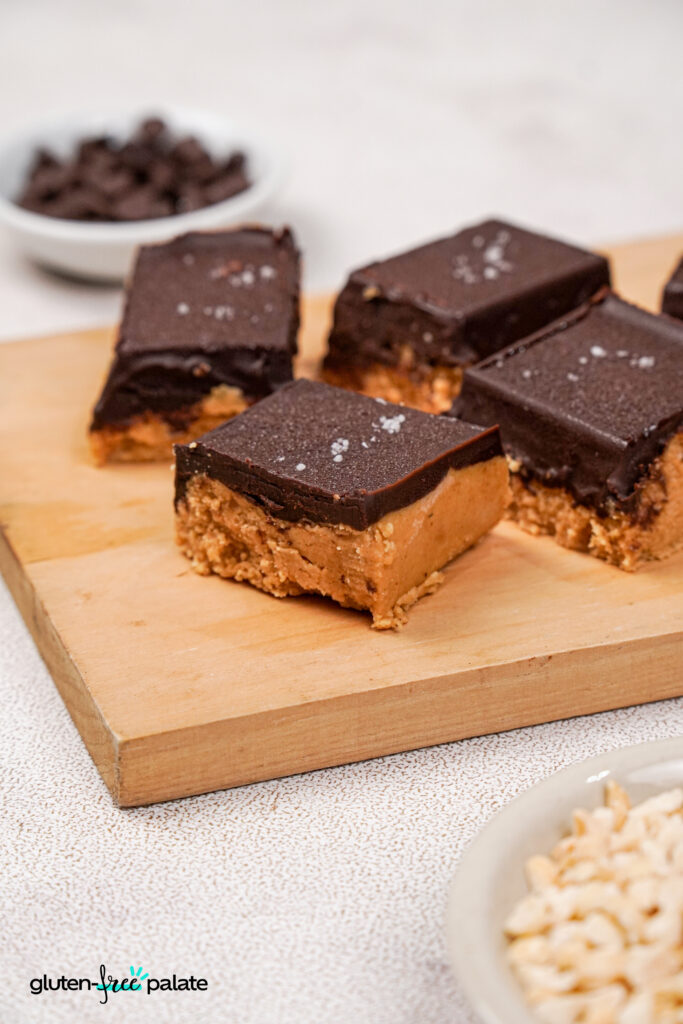 Gluten-free peanut butter bars on a wooden board, with small bowls of chocolate chips and chopped peanuts placed nearby on a light countertop.