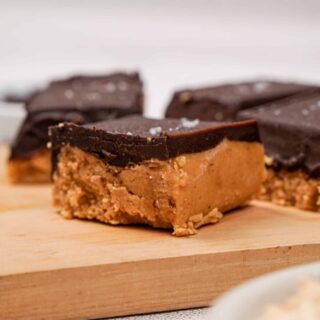 Close-up of thick gluten-free peanut butter bars with a smooth chocolate topping and a sprinkle of flaky sea salt, served on a wooden board with bowls of chocolate chips and chopped peanuts in the background.