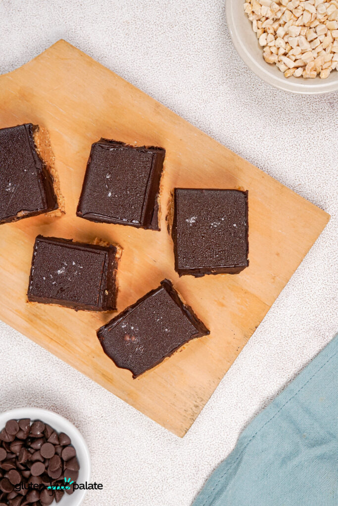 Overhead view of five gluten-free peanut butter bars neatly arranged on a wooden board, each topped with flaky sea salt, with bowls of chocolate chips and chopped peanuts nearby.