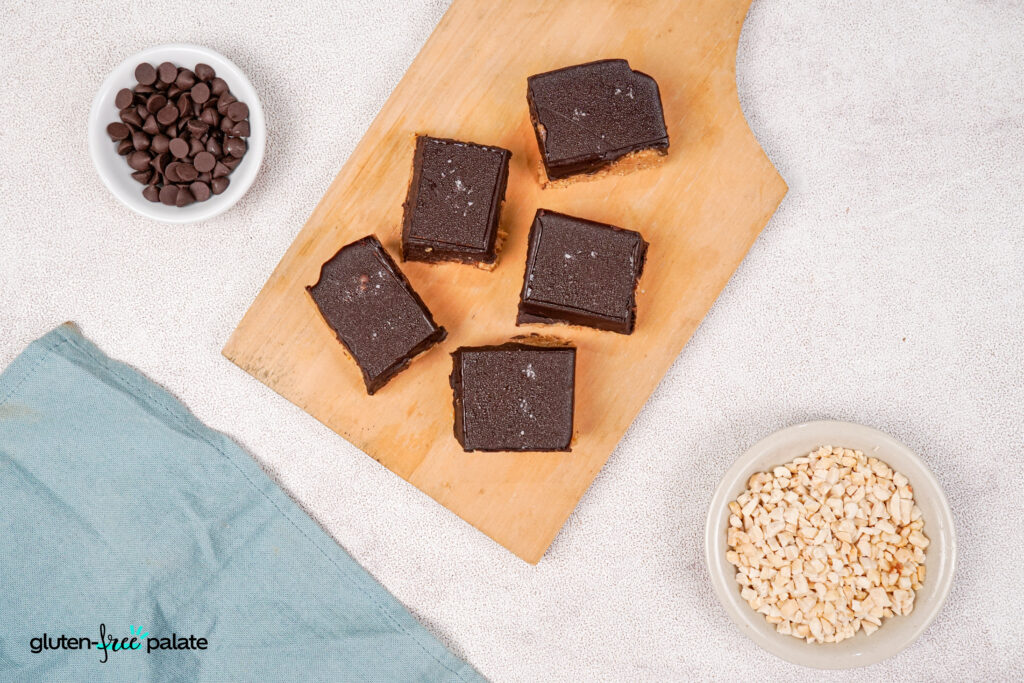Chocolate-topped gluten-free peanut butter bars on a wooden board, showing a rich peanut butter layer with a crunchy base, surrounded by ingredients in small bowls.