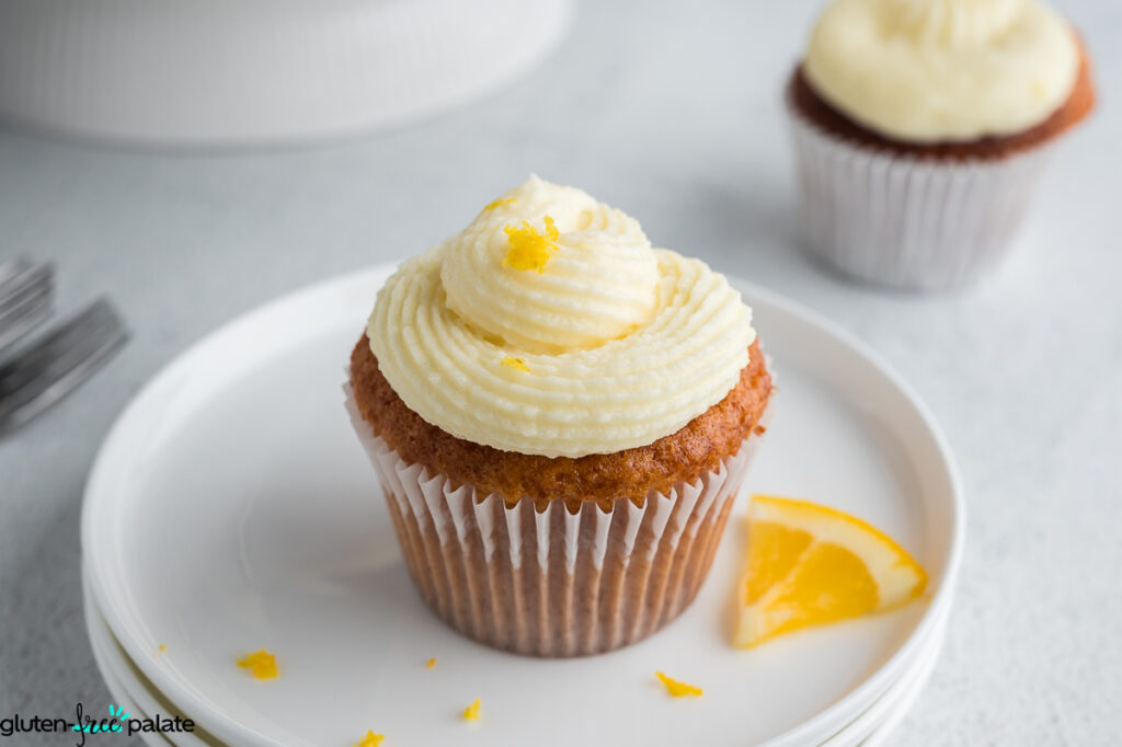 Angled top view of a gluten-free lemon elderflower cupcake topped with swirled frosting and fresh lemon zest, accompanied by a lemon slice.
