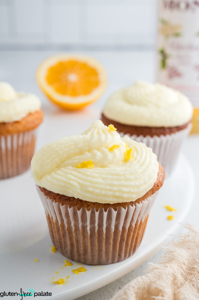 Front view of three gluten-free lemon elderflower cupcakes topped with creamy frosting and lemon zest, displayed on a white serving plate with fresh lemon halves in the background.