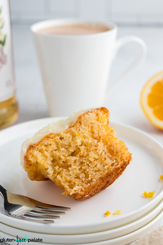 Half-eaten gluten-free lemon elderflower cupcake with soft texture on a plate, served alongside a cup of tea and fresh lemon slices in the background.