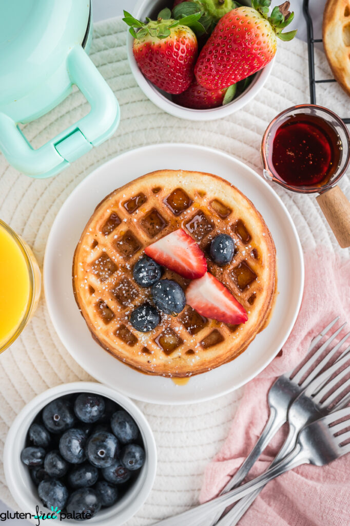 Single mochi waffle on a white plate topped with strawberries, blueberries, powdered sugar, and maple syrup.