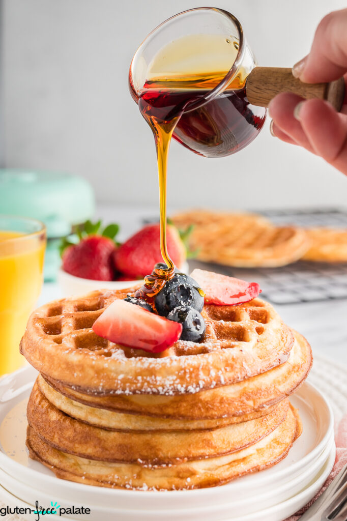 Hand pouring maple syrup onto a stack of mochi waffles topped with strawberries and blueberries.