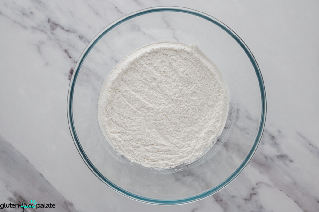 Glass bowl filled with dry ingredients, including sweet rice flour, tapioca starch, sugar, baking powder, and salt.
