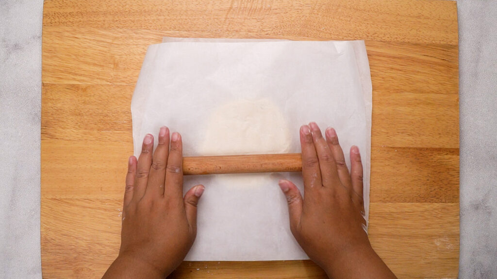 Dough being rolled out between two sheets of parchment paper with a rolling pin.