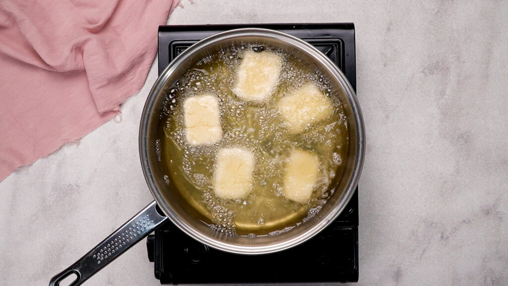 Beignet dough squares frying in bubbling hot oil in a pan.