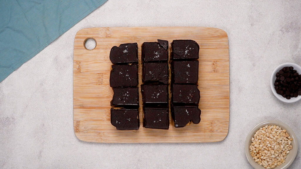 Peanut butter bars cut into neat squares on a wooden cutting board, each topped with a sprinkle of flaky sea salt.