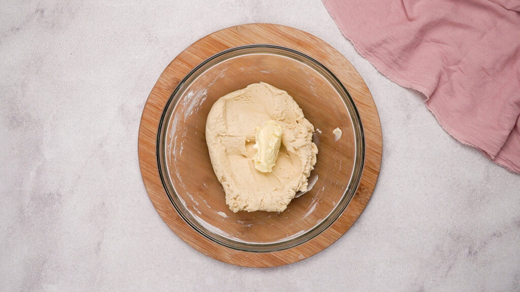 Beignet dough in a glass bowl with softened butter placed on top, ready to be mixed in.
