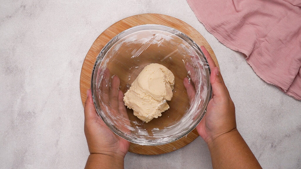 Smooth beignet dough covered with plastic wrap in a glass bowl, set aside for rising.