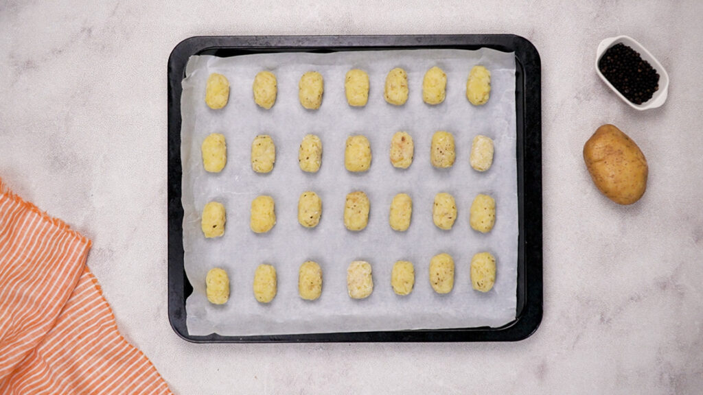 Raw tater tots shaped and lined up on a baking tray with parchment paper.