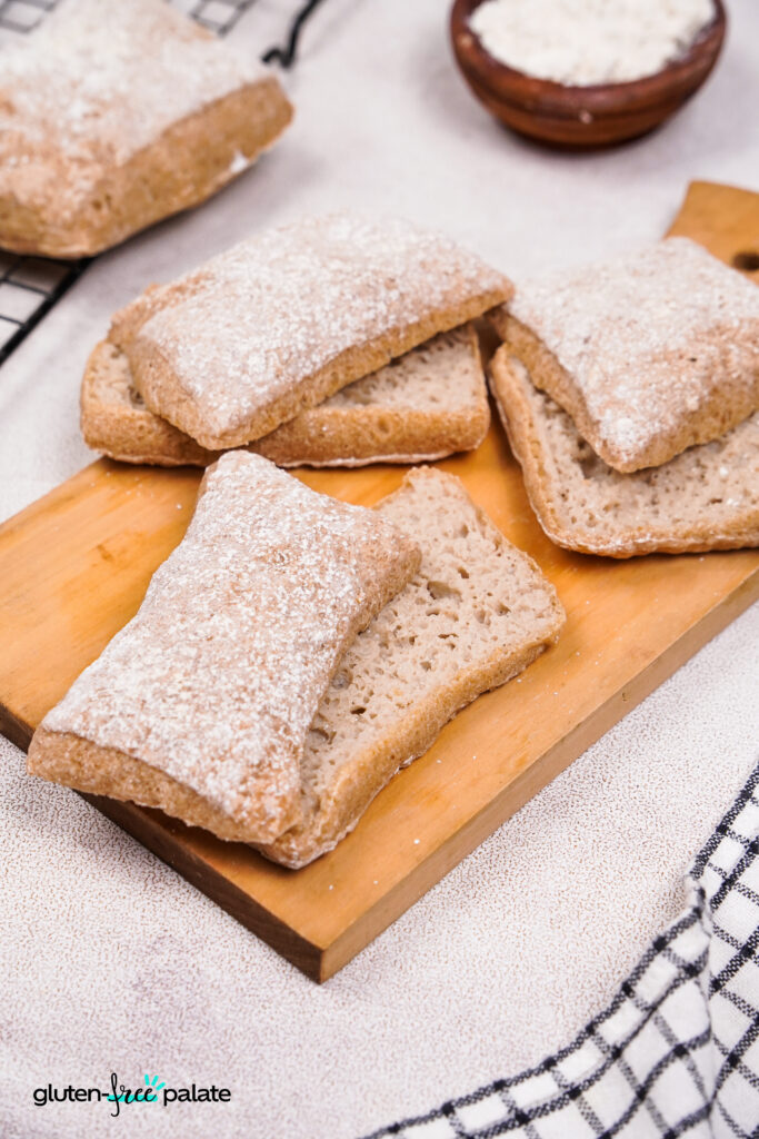 A group of gluten-free ciabatta rolls on a wooden board, lightly dusted with flour, ready to serve.