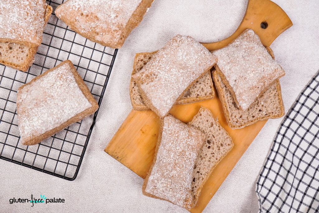 Overhead view of gluten-free ciabatta rolls on a wooden board and cooling rack, with a rustic flour-dusted finish.