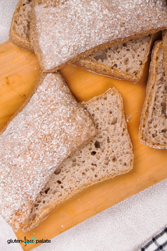 A sliced gluten-free ciabatta roll on a wooden board, showing its airy crumb and dusting of flour on top.